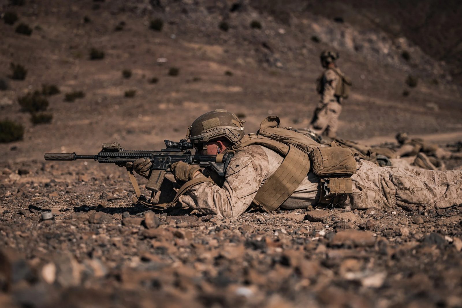 A Marine in the prone position aiming a rifle, with another Marine standing behind
